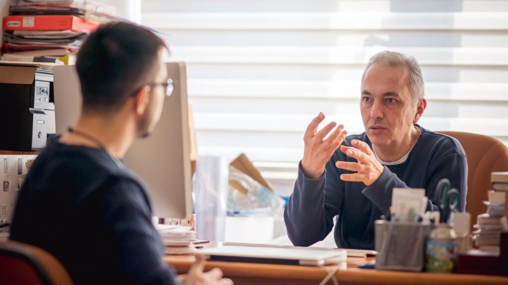 Academic Advising Support Services. Man speaks with another man behind desk in lit room.